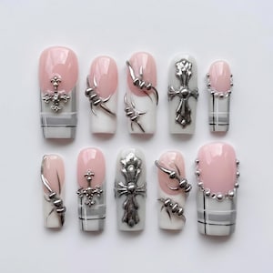 Puede incluir: Un juego de diez uñas postizas con base rosa claro y blanca con un patrón de cuadros gris. Las uñas están decoradas con dijes de metal plateado, que incluyen cruces ornamentadas, diseños de alambre de púas y pequeñas esferas metálicas.