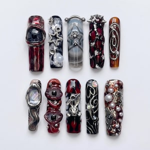May include: A set of ten press-on nails featuring diverse 3D designs including a realistic eye, silver star charms, metallic cross emblems, spikes, a rabbit figurine, and white pearls. The color palette consists of deep red, black, and silver.