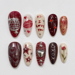 May include: A collection of ten artificial nails with diverse designs. The nails showcase colours like red, white, and brown. Designs include a globe, lips, hearts, stars, and the text "I Love You" and "XOXO".