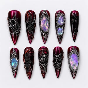 Könnte beinhalten: Ein Set aus zehn langen, spitz zulaufenden Kunstnägeln. Jeder Nagel hat eine tiefrote Basis mit aufwendigen silbernen und schwarzen Designs. Einige Nägel haben schillernde Steinakzente, andere strukturierte Muster.