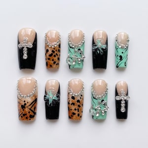 Könnte beinhalten: Ein Set aus zehn künstlichen Fingernägeln mit nudefarbener Basis und verschiedenen Designs, darunter schwarzer Lack, Leopardenmuster und mintgrüne Wirbel. Die Nägel sind mit silberfarbenen Strasssteinen, Sternen und Kreuzen verziert.