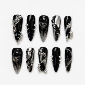 Könnte beinhalten: Ein Set aus zehn stilettoförmigen Press-on-Nägeln mit glänzend schwarzer Basis. Die Designs umfassen silberne Metallic-Akzente, dreidimensionale Totenkopfmotive, spitze Dornen, filigrane drahtartige Wirbel, kleine Silberkugeln und eine feine Kette.