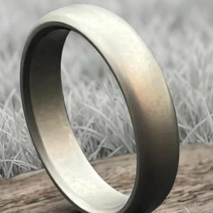May include: A silver-colored ring with a rounded band. The ring is displayed on a wooden surface with a blurred background of grass. The ring's smooth surface reflects light, highlighting its metallic sheen. A simple, elegant piece of jewelry.
