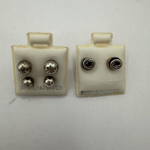 May include: Two white display cards holding sterling silver earrings. The left card features two pairs of round silver studs, one pair with star engravings. The right card holds one pair of silver spiral stud earrings. Each card is labeled STERLING SILVER.