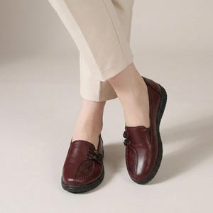 Può includere: Un paio di mocassini in pelle bordeaux scuro con finitura liscia, punta arrotondata e due decorazioni a nodo sul lato della tomaia. Le scarpe hanno un profilo basso con suola nera testurizzata e sono indossate con pantaloni color beige chiaro.