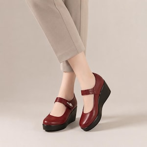 Pode incluir: Um par de sapatos de cunha tipo Mary Jane em vermelho escuro com biqueira arredondada, uma tira sobre o peito do pé com um pequeno botão decorativo prateado e uma cunha preta. Os sapatos são usados com calças de alfaiataria bege claro.