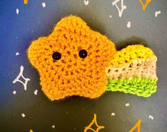 May include: A handmade, crocheted star with black button eyes and a striped tail. The star is a vibrant orange color, and the tail features green, beige, and white stripes. The background is dark blue with drawn stars.