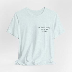 Könnte beinhalten: Hellblaues T-Shirt mit dem schwarzen Text "professionell satt nach 3 Bissen". Das Shirt hat kurze Ärmel und einen Rundhalsausschnitt. Der Stoff scheint weich und meliert zu sein.