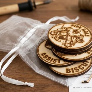 May include: A stack of circular wooden tokens featuring laser-engraved ice hockey imagery and text including Five Hole, Assist, and Biscuit. The tokens are accompanied by a white sheer drawstring pouch and a small wooden hockey stick cutout.