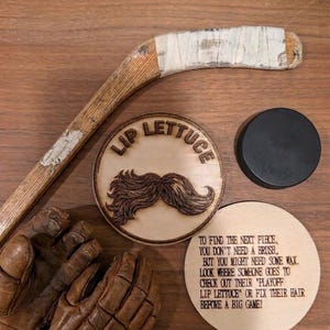 May include: A wooden hockey stick with white tape, a pair of brown leather hockey gloves, a black hockey puck, and two circular wooden plaques. One plaque features a wood-burned moustache and the text "LIP LETTUCE," while the other contains a printed riddle.