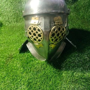 Könnte beinhalten: Ein silberfarbener Gladiator-Helm aus Metall mit einer abgerundeten Kuppel und einem Gesichtsschutz mit zwei kreisförmigen, messingfarbenen Augengittern. Die Gitter sind mit kleinen Messingscharnieren befestigt und der Helm hat einen Nackenschutz.