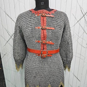 May include: A chainmail tunic featuring a silver-toned interlocking metal ring construction with three-quarter length sleeves. The garment includes a vertical orange leather strap with brass-toned buckles and a matching orange leather waist belt.