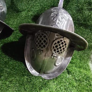 Könnte beinhalten: Ein silberfarbener Gladiator-Helm aus Metall mit breiter Krempe, einem zentralen Kamm und zwei runden Augenschutzgittern mit Gittermuster. Der Helm hat eine glatte, polierte Oberfläche und eine markante, abgerundete Form.