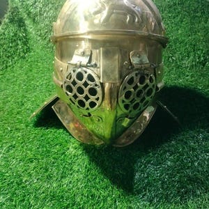 May include: A polished brass gladiator helmet featuring a rounded dome with an embossed animal figure on the top. The front includes two circular, hinged eye guards with a grid pattern and a central nose guard, finished with a wide neck protector.