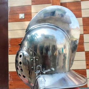 May include: A polished silver-coloured metal helmet featuring a high, curved crest, a rounded dome, and a hinged visor with a series of circular ventilation holes on the side, secured by a metal latch mechanism and a wide neck guard at the base.