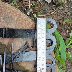 Puede incluir: Bisagras de correa de metal negro con extremos decorativos en espiral y clavos a juego de cabeza cuadrada. Una regla de metal está colocada verticalmente contra las bisagras, mostrando una medida de unos 13 cm de longitud.
