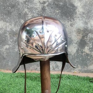 May include: A polished silver-coloured metal helmet featuring a rounded dome and a decorative fan-shaped embossed pattern on the front. The helmet includes a curved brim, visible rivets along the edges, and thin brown leather chin straps hanging down.