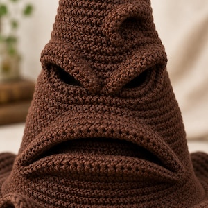 May include: A brown crocheted hat featuring a tall, pointed, conical shape with a wide brim. The hat has a textured surface with visible stitches, including a prominent, folded brow ridge and a wide, horizontal mouth opening near the base.