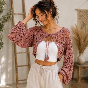 May include: A dusty rose crochet bolero featuring an open-knit pattern, long balloon sleeves, and a front tie closure with two tassels. The garment is worn over a plain white tube top and light-coloured trousers, showcasing the intricate texture.