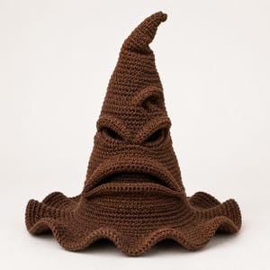 May include: A brown crocheted hat with a tall, pointed, and slightly bent top. The hat features a wide, wavy brim and a textured surface that forms a distinct facial expression with deep, creased folds resembling eyes and a wide, downturned mouth.