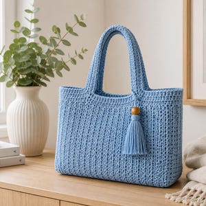 May include: A light blue rectangular crochet tote bag featuring a textured stitch pattern, two sturdy top handles, and a decorative matching tassel accessory attached to one handle with a single round wooden bead at the top of the tassel.