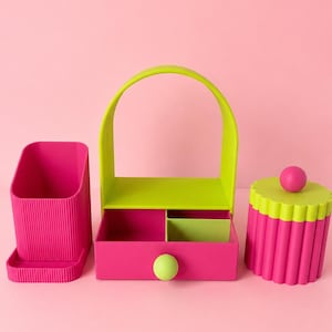 May include: A set of three desk organisers in bright pink and lime green. The left piece is a ribbed pen holder, the centre is a handled caddy with a drawer and lime green knob, and the right piece is a cylindrical container with a scalloped lid.