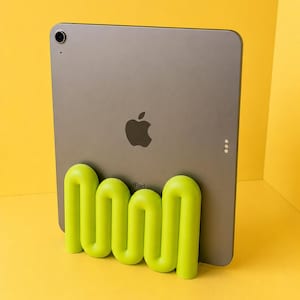 May include: A dark grey tablet with a black Apple logo and the word iPad printed on the back, resting in a bright lime green, wavy, tubular stand. The stand features a continuous, serpentine shape that supports the device in an upright position.