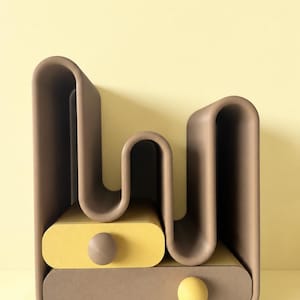 May include: A taupe-coloured storage organiser featuring a wavy, multi-tiered structure with two horizontal drawers. The top drawer is yellow with a taupe knob, and the bottom drawer is taupe with a yellow knob, creating a layered, geometric design.