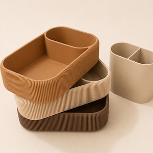May include: A stack of three rectangular storage trays with rounded corners and vertical ribbed exterior walls, coloured in shades of tan, beige, and dark brown, alongside a separate upright oval container with a central divider.