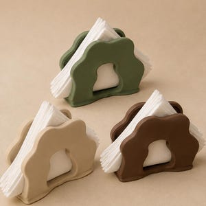 May include: Three napkin holders in olive green, beige, and dark brown, each featuring a wavy, organic-shaped frame with a central cutout. Each holder contains a stack of folded white paper napkins, displayed in a triangular arrangement.