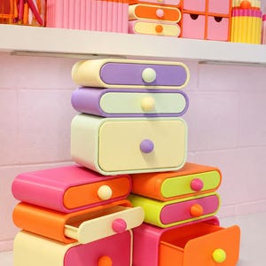 May include: A stack of modular desk organisers featuring rounded rectangular drawers in pastel and vibrant colours including pink, orange, yellow, purple, and lime green. Each drawer has a single round knob in a contrasting colour for easy opening.