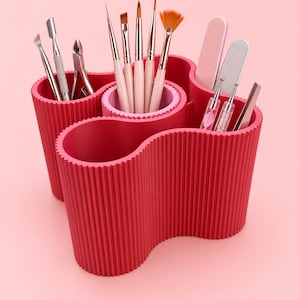 May include: A bright pink, ribbed, multi-compartment organiser holding various metal nail tools, makeup brushes, nail files, and dotting tools. The container features a wavy, organic shape with distinct sections for sorting different beauty implements.