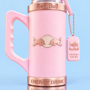 May include: A light pink beverage can holder with a handle, featuring the Red Bull logo in rose gold. The top rim reads WITH MONK FRUIT and the base reads ENERGY DRINK. A small matching rose gold tag hangs from a ball chain on the side.