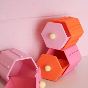 May include: A set of three hexagonal storage containers with drawers. One container is orange with a pink drawer, one is pink with an orange drawer, and one is light pink with a matching light pink drawer. Each drawer features a small yellow knob.