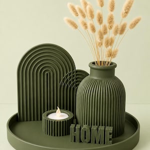 May include: A dark olive green decorative set on a round tray featuring a ribbed vase, a ribbed candle holder with a lit tea light, two arched panels with vertical grooves, and the word HOME in block letters, with beige dried grass stems.
