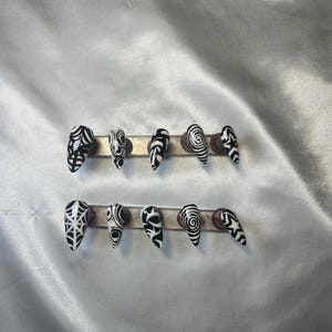 May include: A set of ten black and white press-on nails. Each nail has a unique design, such as spiderwebs, swirls, and geometric patterns. The nails are displayed on a silver bar against a white satin background.