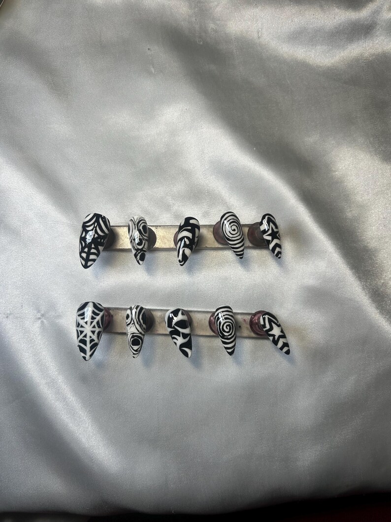 May include: A set of ten black and white press-on nails. Each nail has a unique design, such as spiderwebs, swirls, and geometric patterns. The nails are displayed on a silver bar against a white satin background.