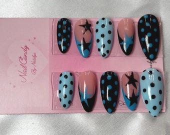 May include: A set of ten oval false nails with diverse designs. Some nails have black polka dots on a light blue base, others feature black stars with blue accents, and some have a pink and black gradient. The packaging reads "Nail Candy By Natalya."