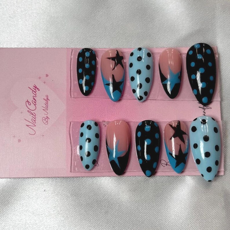 Pode incluir: Um conjunto de dez unhas posti&ccedil;as ovais. As unhas apresentam v&aacute;rios designs, incluindo bolinhas pretas sobre um fundo azul claro, estrelas pretas com detalhes em azul e um gradiente rosa e preto. A embalagem diz "Nail Candy By Natalya."