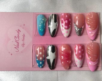 May include: A collection of ten false nails with diverse designs. The nails showcase shades of blue, pink, and silver, with patterns such as polka dots and stars. They are presented on a pink card, which has the text "Nail Candy By Nostalgia."