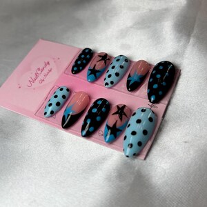 Pode incluir: Um conjunto de dez unhas posti&ccedil;as ovais. As unhas apresentam v&aacute;rios designs, incluindo bolinhas pretas em fundo azul claro, estrelas pretas em fundo rosa e preto e bolinhas pretas em fundo preto. Um cart&atilde;o rosa com o texto "Nail Candy By Nataliya" est&aacute; no fundo.