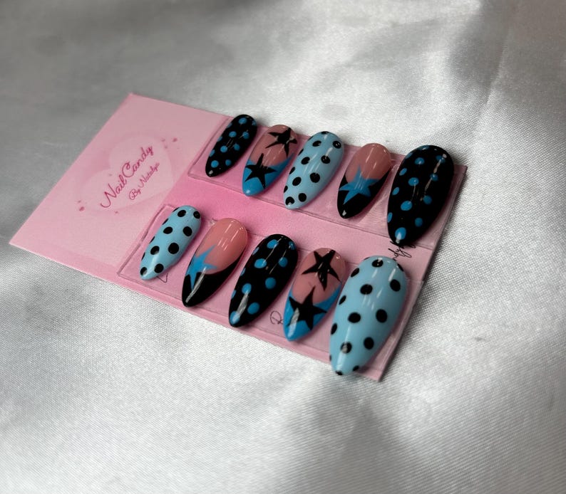 Pode incluir: Um conjunto de dez unhas posti&ccedil;as ovais. As unhas apresentam v&aacute;rios designs, incluindo bolinhas pretas em fundo azul claro, estrelas pretas em fundo rosa e preto e bolinhas pretas em fundo preto. Um cart&atilde;o rosa com o texto "Nail Candy By Nataliya" est&aacute; no fundo.