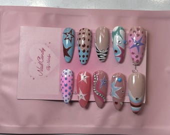May include: A collection of ten false nails with diverse designs, such as stars, polka dots, and abstract patterns. The nails feature blue, pink, and nude tones, some embellished with rhinestones. The Nail Candy logo is displayed on a pink card.