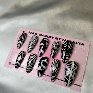 May include: A set of ten long, almond-shaped press-on nails with black and white abstract designs. The designs include geometric patterns, swirls, and web-like motifs. The nails are displayed on a pink card with the text "NAIL CANDY BY NATALYA".