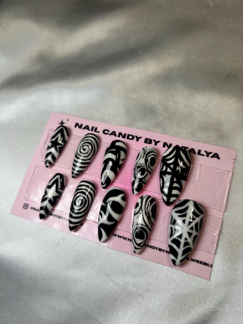 May include: A set of ten long, almond-shaped press-on nails with black and white abstract designs. The designs include geometric patterns, swirls, and web-like motifs. The nails are displayed on a pink card with the text "NAIL CANDY BY NATALYA".