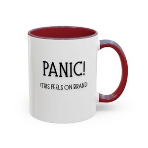 May include: A white ceramic mug with a dark red interior and handle. The front of the mug features black text that reads "PANIC!" in a large font, followed by the smaller text "(THIS FEELS ON BRAND)" centered directly underneath the main word.