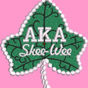May include: A green leaf-shaped emblem with the text "AKA Skee-Wee" in white. The leaf has a white beaded border and black decorative lines. The background is a solid pink color.