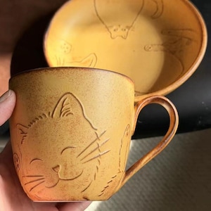 May include: A ceramic mug and saucer set in a warm, earthy tone. The mug features an embossed cat face, while the saucer has cat outlines. The set is designed with a rustic, handcrafted aesthetic, perfect for enjoying coffee or tea.