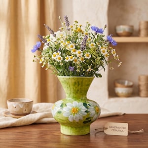 May include: A lime green ceramic vase with a flared rim and a rounded body, featuring hand-painted white daisy motifs with yellow centers. A small rectangular tag attached to the base with twine displays the text HANDPAINTED CERAMICS.
