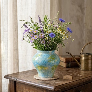 May include: A ceramic vase with a mottled blue, green, and white finish holds a bouquet of wildflowers including purple lavender, blue cornflowers, and white daisies. The vase sits on a round woven mat, accompanied by a small tag labeled Coastal Ceramics.